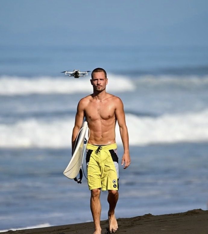 Tony walking up the beach with his surfboard, a drone filming behind him